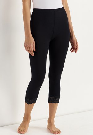 Vrouw draagt zwarte cropped leggings met kanten rand aan de zoom, staat op blote voeten tegen een effen lichte achtergrond.