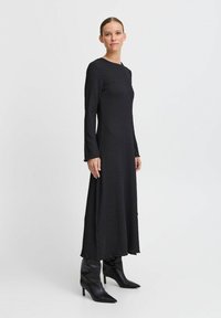 Musta pitkähihaisen mekko, jossa on teksturoitu kangas, löysä istuvuus ja pyöreä pääntie; yhdistetty mustiin polvipitkiin kärjellisen vaelluskengän kanssa.
