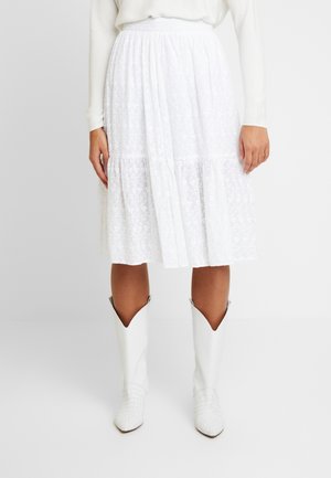 Femme portant une jupe midi blanche à texture florale et des bottes de cow-boy blanches, debout devant un fond blanc uni.