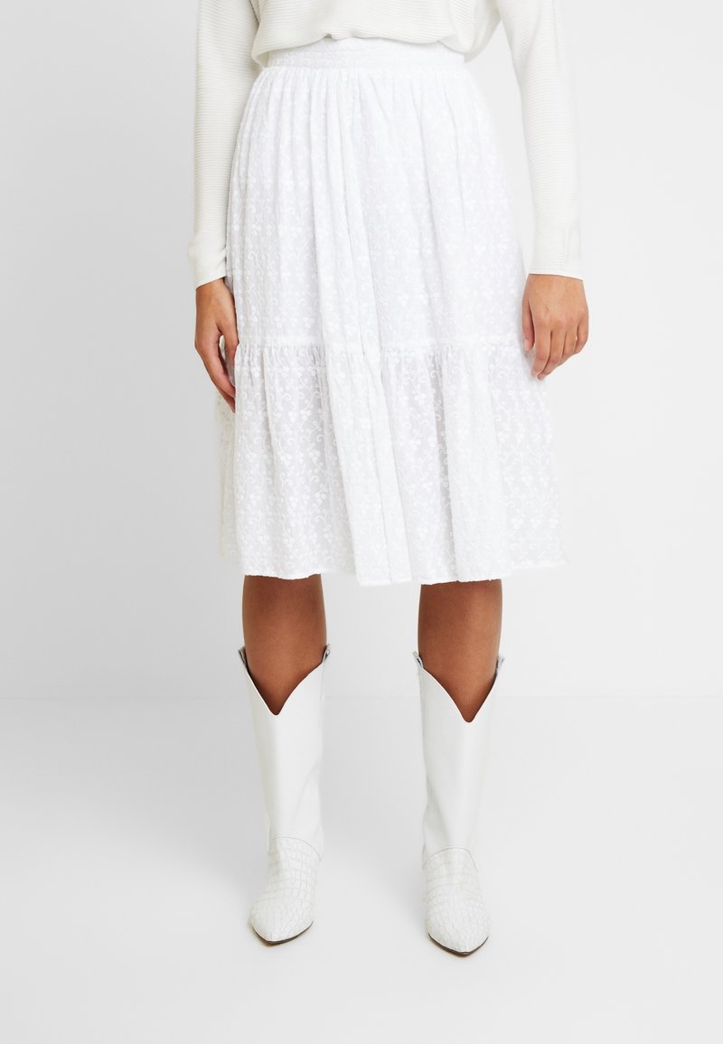 Femme portant une jupe midi blanche à texture florale et des bottes de cow-boy blanches, debout devant un fond blanc uni.