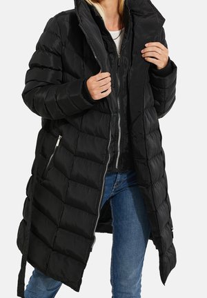 Personne portant un long manteau matelassé noir sur une veste noire zippée et un jean bleu, ajustant le col du manteau.