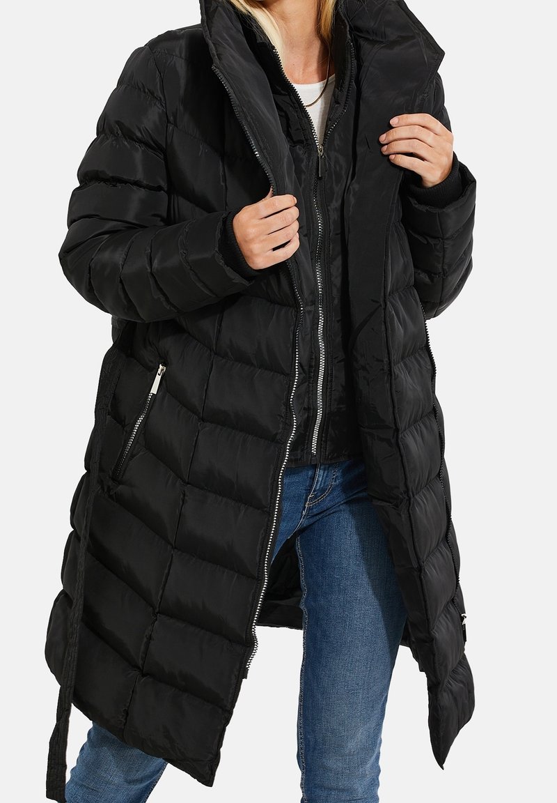 Personne portant un long manteau matelassé noir sur une veste noire zippée et un jean bleu, ajustant le col du manteau.