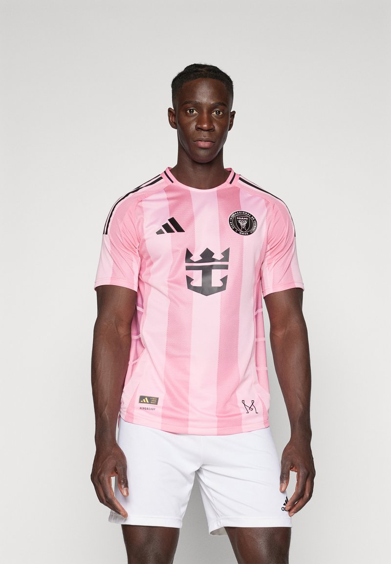 Lyserød fodboldtrøje med sorte detaljer, der har lodrette striber, holdlogo og et krone-design. Matchende med hvide shorts for et komplet look.