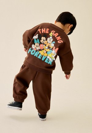 Child wearing brown sweatshirt and pants with colorful Disney characters and the text "The Gang Forever" on the back, mid-step against beige background.