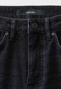 Jean en denim noir avec une texture lisse, présentant une fermeture à bouton et une étiquette à la taille portant l'inscription "un jour." Détails de couture minimaux.