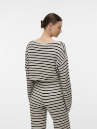 Γυναίκα με κότσο στα μαλλιά, που φοράει μπεζ και μαύρο πουλόβερ με οριζόντιες ρίγες και ταιριαστό παντελ όνι, στέκεται και κοιτάζει πάνω από τον ώμο της.