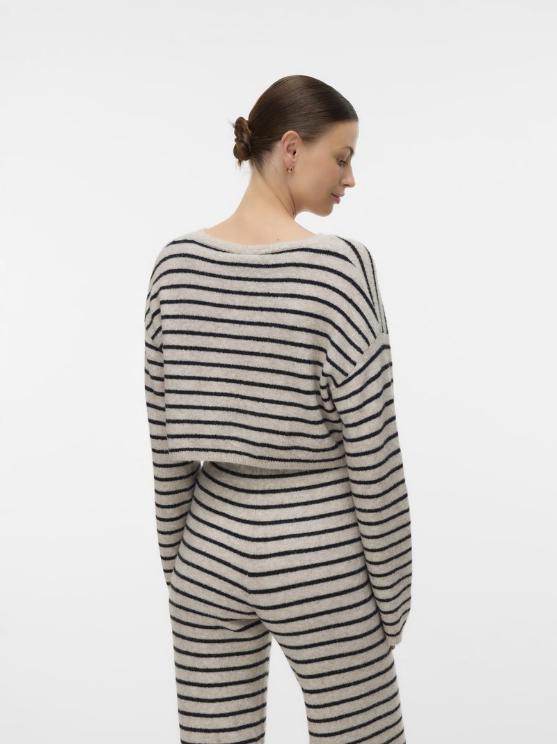 Γυναίκα με κότσο στα μαλλιά, που φοράει μπεζ και μαύρο πουλόβερ με οριζόντιες ρίγες και ταιριαστό παντελόνι, στέκεται και κοιτάζει πάνω από τον ώμο της.