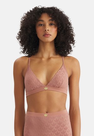 Frau mit lockigem Haar, die einen passenden rosafarbenen Bralette und hoch taillierte Unterteile mit dezentem Blattmuster und Adidas-Logos trägt.