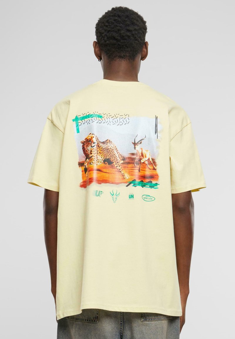 T-shirt en coton jaune avec un imprimé graphique d'un léopard et d'un antelope au dos, agrémenté d'accents verts et rouges et de divers détails de logo.