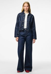 Traper jakna i široke traperice, obje u tamnom indigu. Jakna ima skraćeni kroj s metalnim gumbima, a traperice imaju visok struk.