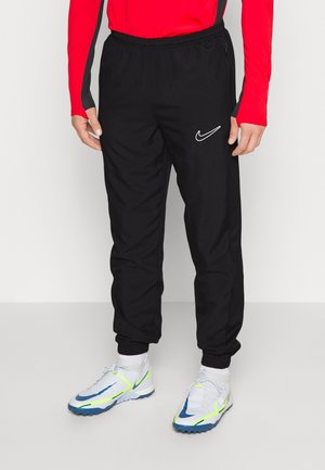 Personne portant un pantalon de survêtement Nike noir, des chaussures de sport blanches avec des détails bleus et jaune néon, et un haut rouge à manches longues, debout devant un fond uni.