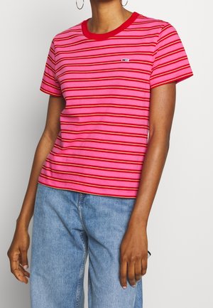 Person iført en pink og rød stribet kortærmet skjorte med et lille logo på brystet og lyseblå jeans.