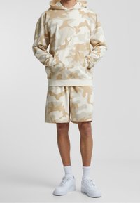 Haut à capuche camouflage beige avec une poche avant et un short assorti. Assorti avec des baskets blanches. Tissu doux, coupe décontractée et sans motifs visibles.