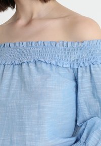 Off-shoulder blus i ljusblå bomull med volang  vid halsringningen, lätt och luftig textur samt subtilt vävt mönster. Avslappnad passform med korta ärmar.