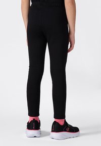 Persona vestida con leggins negros, calcetines rosas y zapatillas deportivas negras con suelas blancas, de pie contra un fondo claro y liso.