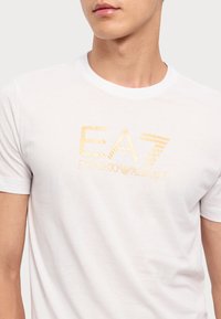 Hvit bomulls t-skjorte med et gull EA7 Emporio Armani-logo over brystet. Korte ermer og klassisk rund hals.