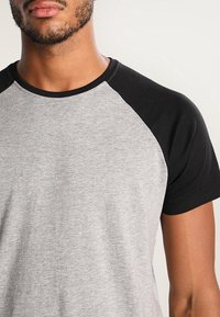 Szara bawełniana koszulka z czarnymi rękawami raglanowymi, z okrągłym dekoltem i gładką fakturą. Warstwowy kontrast kolorów wzdłuż ramion.