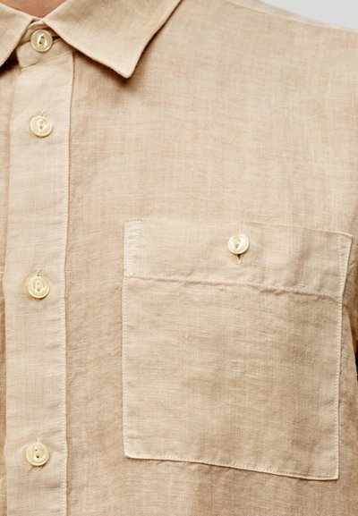Μπεζ λινό πουκάμισο με γιακά, έξι κουμπιά και τσέπη στο στήθος. Το ύφασμα έχει υφή και σχεδιασμό casual.