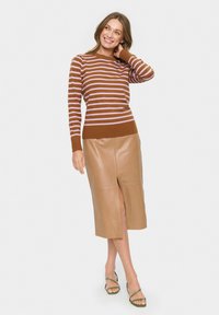 Frau, die lächelt, trägt einen gestreiften Pullover in Braun und Rosa, einen beigen Leder-Midi-Rock mit Schlitz vorne und grüne Sandalen mit Riemchen, steht vor einem weißen Hintergrund.