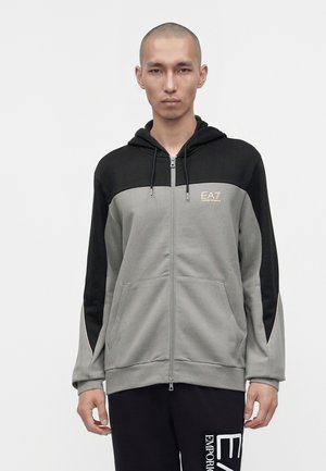 Mann trägt einen grau-schwarzen Zip-Hoodie mit EA7 Emporio Armani Logo auf der Brust und schwarze Hose mit großem weißen Text am Bein.