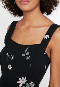 Robe noire à imprimé floral, dotée de larges bretelles, d’un décolleté carré et d’ornements de fleurs roses et de feuilles vertes.