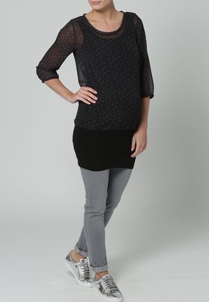 Femme en blouse noire transparente à pois, jean skinny gris et baskets argentées, debout avec une main sur la hanche sur un fond clair uni.