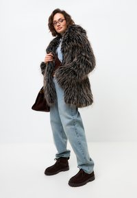 Pardoseală din blană sintetică, de culoare neagră și maro, cu un croi oversized, asortată cu blugi din denim de un albastru deschis și ghete din suede maro închis.
