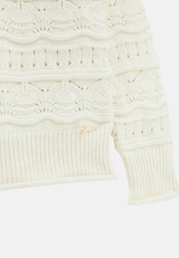 Suéter blanco de punto con intrincados patrones en forma de ola, un dobladillo acanalado y un pequeño acento de logo dorado. Textura suave y diseño ajustado.