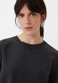 Schwarzer Sweatshirt mit rundem Halsausschnitt, glatter Textur und dezenten Nähten. Das Modell trägt silberne Creolen. Einfarbiger Hintergrund.