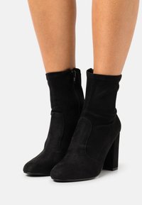 Bottes cheville en suède noir avec une coupe ajustée, fermeture éclair latérale, bout pointu et talon haut épais. Texture lisse avec des détails de couture subtils.