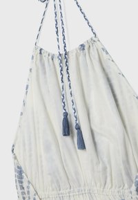 Tissu blanc avec un motif floral bleu, doté de cordons bleus tressés se terminant par des pompons, attachés à un col froncé.