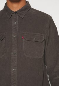 Close-up van een persoon die een donkerbruine corduroy overhemd met knopen draagt, met twee borstzakken en een rood label op de rechterzak.