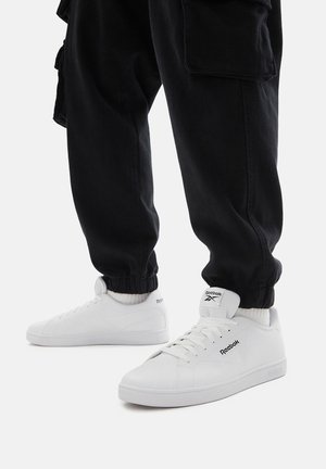 Osoba w czarnych spodniach typu cargo i białych trampkach marki Reebok stojąca na białym tle.