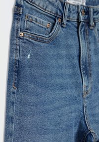 Vaqueros de mezclilla azul de tiro alto con una textura ligeramente desgastada, que cuentan con un pequeño bolsillo frontal, remaches en tono cobre y detalles de costura.