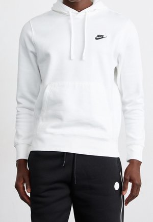 Osoba ubrana w białą bluzę Nike z kapturem i przednią kieszenią oraz czarne spodnie z białym paskiem po boku i okrągłym logo na udzie.