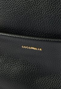 Cartera de piel negra texturizada con borde liso, con un logo dorado "Coccinelle" en el frente. La superficie muestra un patrón de grano fino.