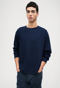 Sötétkék, hosszú ujjú pulóver, texturált anyagból, kerek nyakkivágással és laza szabással. A modellen sötét kordbársony nadrág van.