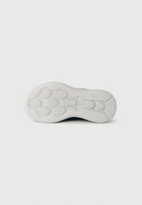 Semelle d'une chaussure de sport blanche avec des motifs de grip texturés et un logo en relief au centre, sur un fond blanc uni.
