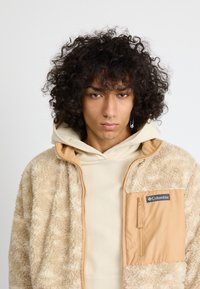 Person mit lockigem Haar, die eine beige Fleecejacke und einen cremefarbenen Hoodie trägt, mit Nasenpiercing und einem langen Ohrring im linken Ohr.