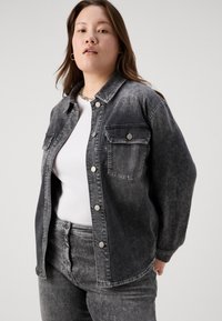Siva denim srajca z gumbi, z dvema žepoma na prsih, srebrnimi dodatki in sproščenim krojem, združena z belo majico in ustreznimi jeans hlačami.