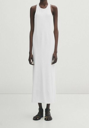 Lange persoon draagt een mouwloze witte jurk tot aan de enkels, gecombineerd met zwarte sandalen met open tenen, staand tegen een effen lichte achtergrond.