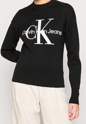 Persona que lleva un suéter negro de Calvin Klein jeans con un gran logo blanco y beige, combinado con pantalones sueltos de color claro, de pie con un brazo doblado.