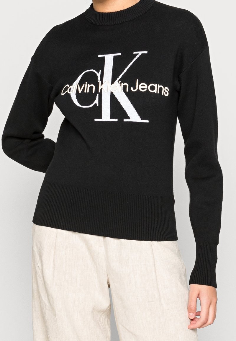 Personne portant un pull Calvin Klein jeans noir avec un grand logo blanc et beige, associé à un pantalon ample de couleur claire, debout avec un bras plié.