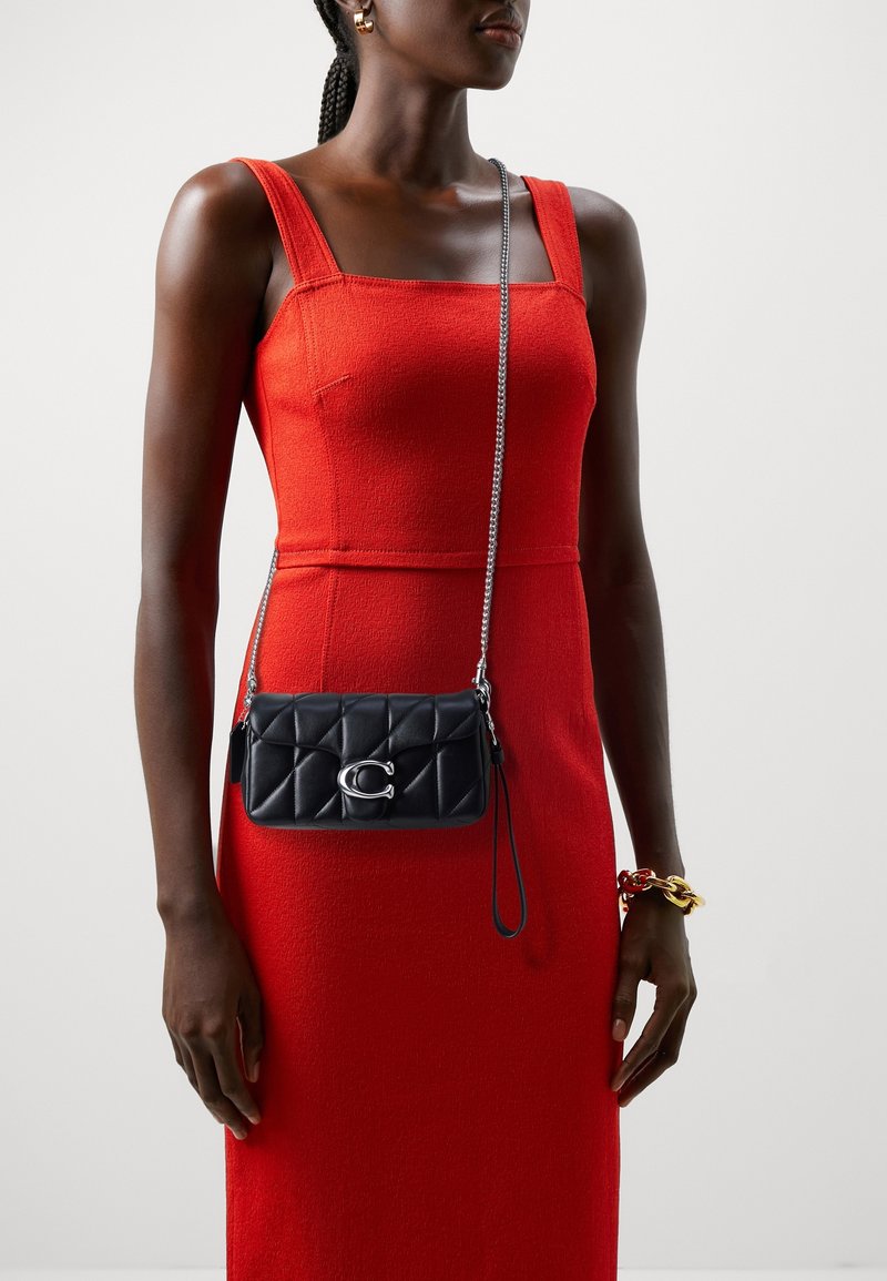 Femme portant une robe rouge sans manches et un bracelet doré, tenant un petit sac bandoulière noir matelassé avec une chaîne argentée et un détail de logo.