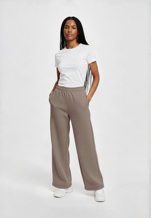 Jeune femme aux longues tresses portant une chemise blanche à manches courtes, un pantalon large marron et des baskets blanches, debout les mains dans les poches.