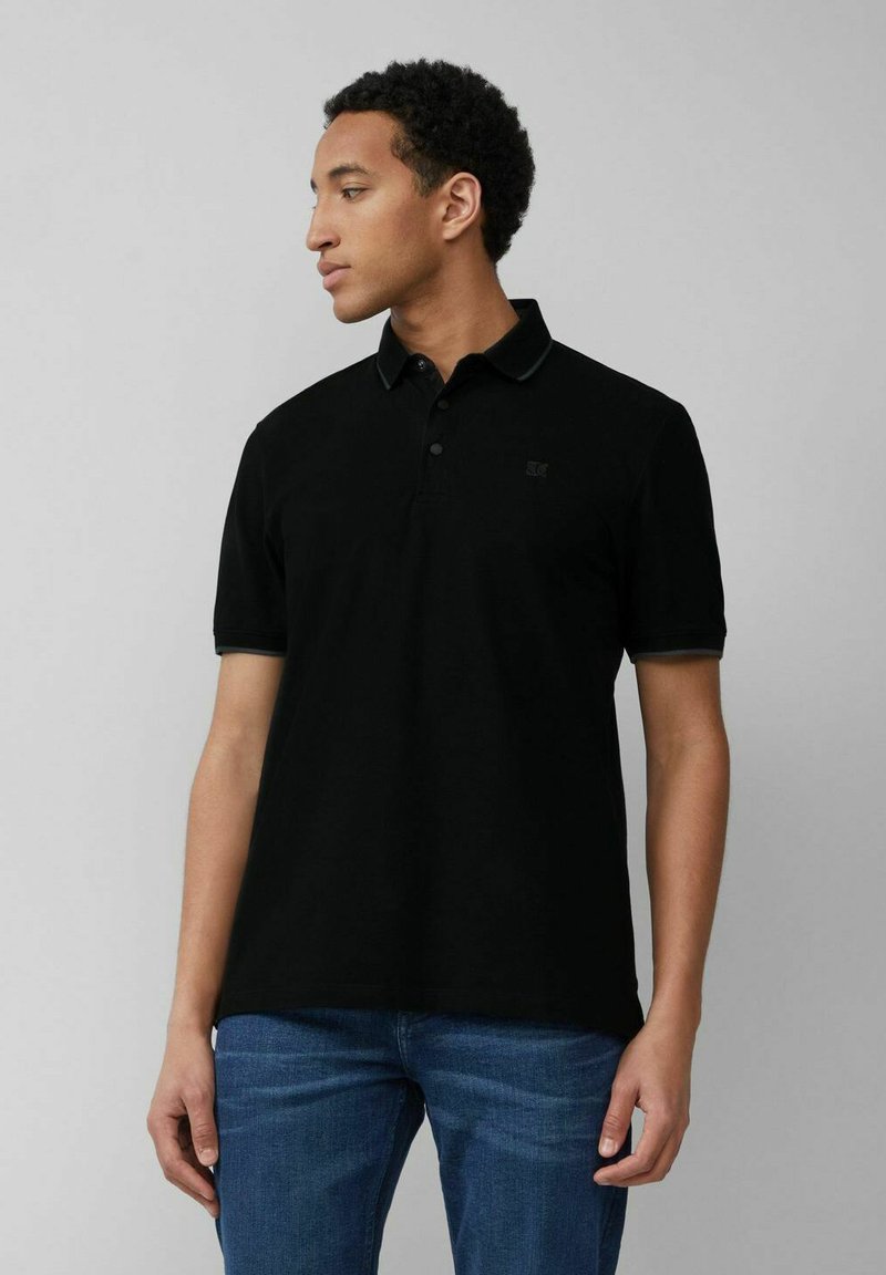 Schwarzes Polo-Shirt mit einer soliden Textur, kurzen Ärmeln, einem Kragen und einer Knopfleiste mit drei Knöpfen. Verfügt über ein dezentes Logo auf der Brust. Getragen mit blauen Jeans.
