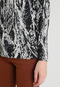 Czarny i biały top z abstrakcyjnym wzorem, z długimi rękawami i luźnym krojem, zestawiony z brązowymi dopasowanymi spodniami.