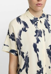 Kurzärmelige Bluse in Creme mit einem abstrahierten navyblauen Blumenmuster, mit spitzem Kragen und Knopfverschluss, aus einem glatten Stoff gefertigt.