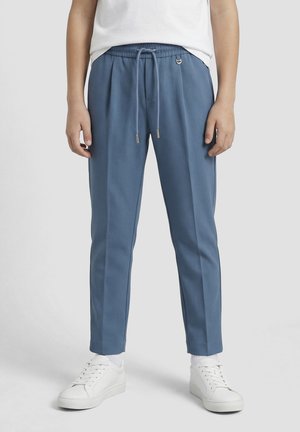 Osoba koja nosi plave uske hlače s vezicom u struku, bijele čarape, bijele tenisice i bijelu košulju, stoji na jednobojnoj pozadini.