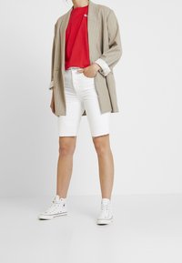 Beige stribet blazer over en rød t-shirt, parret med hvide denimshorts og hvide højhælede sneakers. Ensfarvet baggrund.